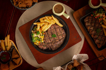 Flatlay angle grilled sirloin steak or tenderloin steak with fries and vegetables. Served on around black hot plate with mushroom sauce. Perfect for advertising or commercial purpose. Top view angle