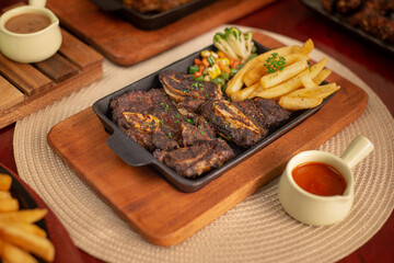 Grilled bbq beef ribs with fries and vegetables. Served on black hot plate with mushroom and bbq sauce. Landscape angle on wooden table. 45 degree angle camera. Perfectly for advertising restaurant