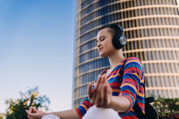 Calm young woman sits cross-legged downtown, embracing sound meditation with closed eyes, enjoying...