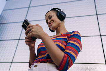 Smiling young woman in colorful striped shirt using smartphone while wearing headphones, enjoying a digital moment in front of a modern metallic architectural wall