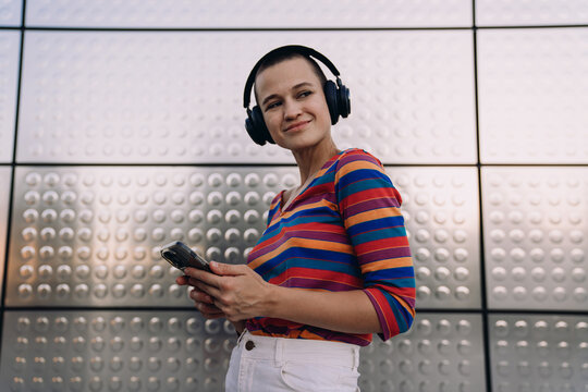 Trendy young woman with buzzcut and headphones holding smartphone, smiling and looking away while enjoying her playlist against a futuristic urban backdrop