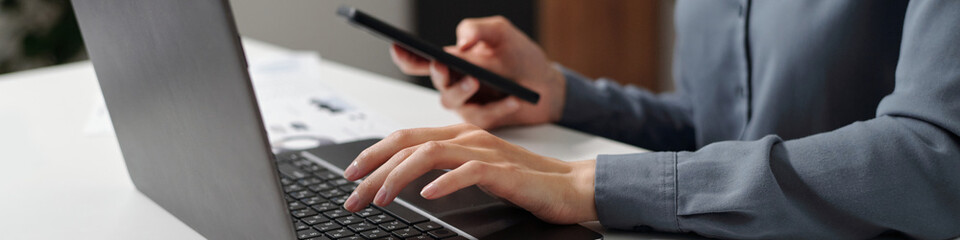 Header image of female hands typing on laptop keyboard and using smartphone in office workplace setting copy space