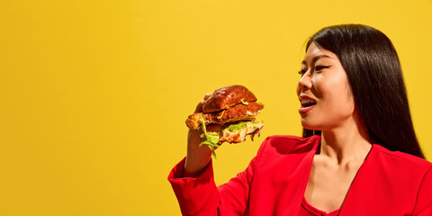 Side view of stylish young woman in red jacket preparing to bite giant burger against bright yellow background. Concept of fast food, pop art, human emotions. Banner. Copy space for ad