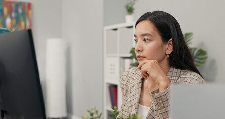 A focused pretty woman with long brown hair wearing a checkered jacket sits at desk in office in...