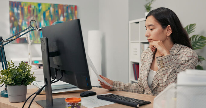 An attractive, experienced company accountant sits in office in front of a computer checking statistical data employee salary charts comparing paper diagrams with electronic ones financial analyst