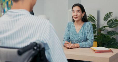 Fototapeta premium In a professional office setting, a wheelchair user in a shirt sits across from a beautiful Asian-Korean woman during a job interview. She listens carefully, taking notes as he answers her questions.