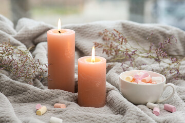 Burning candles and cup with marshmallow on plaid near window