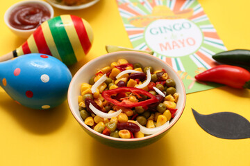 Bowl with traditional Mexican corn salad, maracas and Cinco de Mayo greeting card on yellow background, closeup
