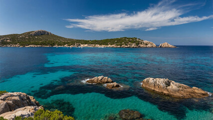 Fototapeta premium Turquoise and deep blue water meets a rocky shoreline with green vegetation under a bright blue sky with fluffy white clouds