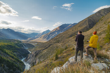 Two photographers with cameras are standing on a slope in bright clothes and taking pictures against the background of snowy mountains, forests and a river that flows through the Altai gorge.