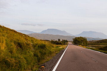 Golden sunset over calm lakes and clear water reflections in The Trossachs and Loch Lomond. Long winding roads cross green hills and mountains in a peaceful, awe-inspiring natural landscape. Exploring