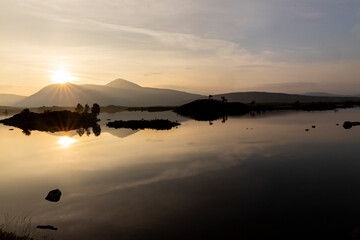 Golden sunset over calm lakes and clear water reflections in The Trossachs and Loch Lomond. Long winding roads cross green hills and mountains in a peaceful, awe-inspiring natural landscape. Exploring