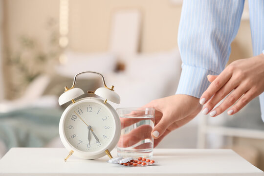 Woman taking pills and glass of water from bedside table with alarm clock in bedroom, closeup