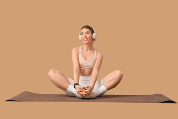 Sporty young woman with headphones sitting on yoga mat against beige background