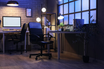 Interior of dark office with desks, shelf unit and glowing lamps at night