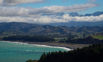 New Zealand nature landscape panorama, turquoise blue ocean, black sand beach, green rolling hills and snow covered mountains seen from Kaikoura