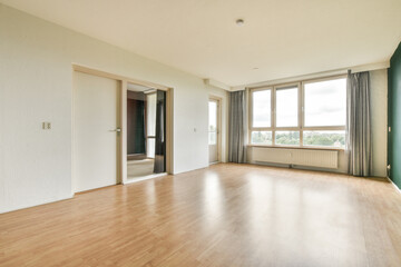 A bright, spacious room featuring large windows that allow natural light to flood in, highlighting the wooden flooring and minimalist design.