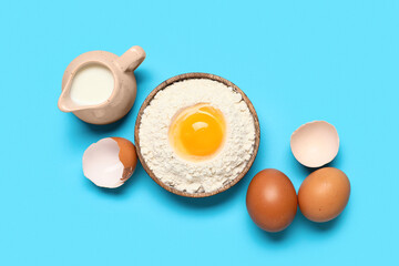 Plate with flour, raw eggs and pitcher of milk on color background