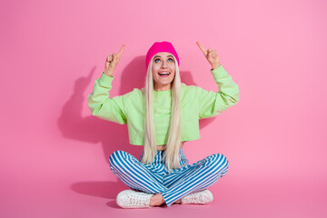 Stylish young woman in vibrant fashion sporting a pink beanie and green sweatshirt, sitting on colorful striped pants