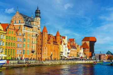 Charming historic architecture lines the waterfront of Gdansk under a clear blue sky. The river is bustling with boats, offering a vibrant scene with the city's iconic brick buildings