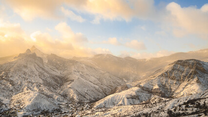 Snow-covered high rocky and rolling mountains in the golden haze light of sunset. A breathtaking winter landscape. Low-hanging clouds. Snowy mountains at sunrise in winter. Nature inspired banner.