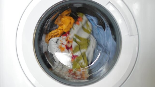 Rotating washing machine drum tumbling colorful wet garments, splashing water and detergent during laundry cleaning process