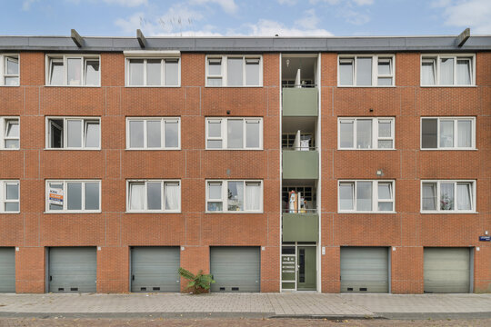 A contemporary multi-story apartment building featuring a brown brick facade and multiple garage doors underneath. Windows are abundant, providing ample light. - Powered by Adobe