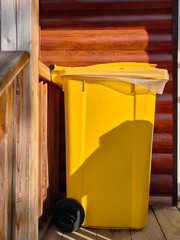 bright yellow trash bin stands beside wooden railing in sunlit area. concept waste disposal and environmental care. contrasting colors and textures. close up.