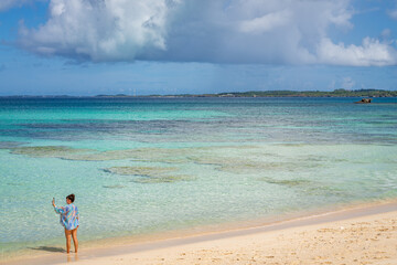 Woman takes selfie on Okinawa beach, capturing the turquoise water and sunny skies..