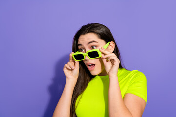 Young woman in neon green outfit adjusts sunglasses with a surprised expression against a vibrant purple background