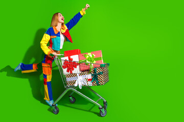 Excited young woman in colorful clothing celebrating shopping success with a cart full of Christmas...