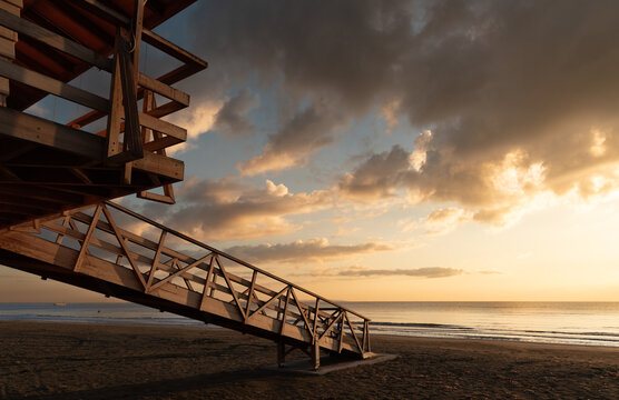Wooden lifeguard tower overlooking sandy beach at sunrise with cloudy sky