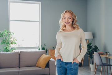 Relaxed young woman with blonde hair standing in a bright, modern living room wearing casual...