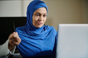Middle Eastern Woman Focuses on Online Work at Her Desk in Blue Hijab