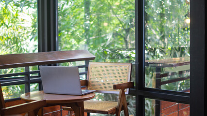 A laptop is placed on a wooden table in a cafe. The view behind is a glass window with a refreshing view of trees, suggesting remote work.