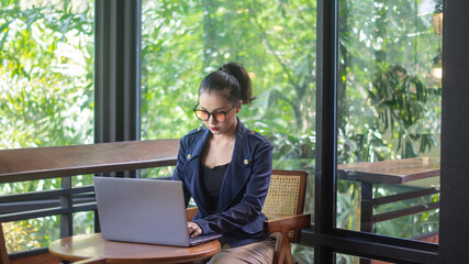 Businesswoman wearing glasses working on laptop in cafe, intently, working remote. A woman works on a computer in a coffee shop with a large mirror and plants.