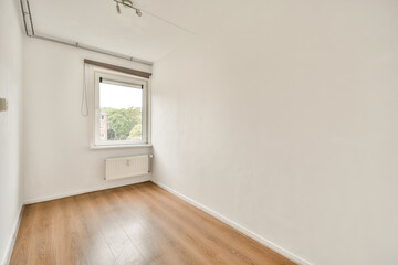 A bright, minimalist room showcasing white walls and a wooden floor. The window allows natural light, enhancing the spacious feel.