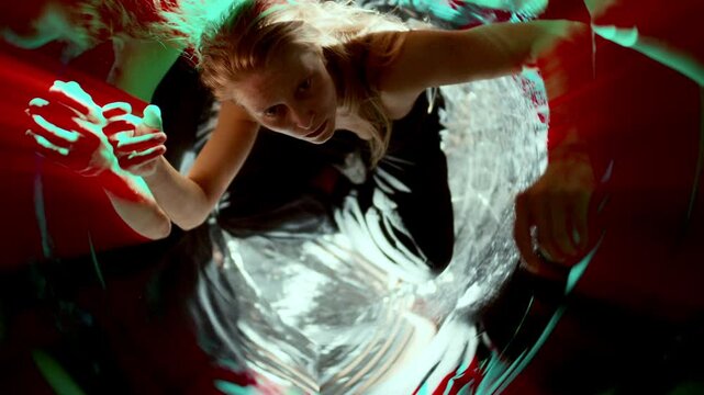 Young woman sitting on shiny floor, raising hands and looking upward, performing inside distorted mirrored space filled with colorful light streaks, High angle view