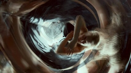 Young female performer moving arms fluidly and gazing around inside mirrored room filled with swirling reflections and immersive light distortions. High angle view