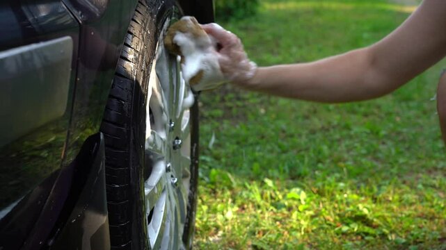 Mans hands wash car wheel with foam and sponge on lawn with green grass near pond or lake. Concept of washing car in prohibited place in nature. Pollution of environment