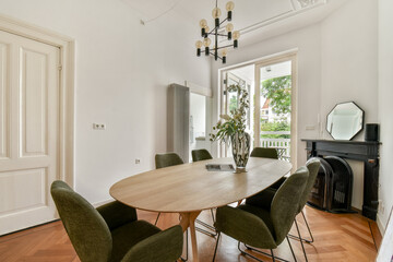 A stylish dining room featuring a round wooden table surrounded by green chairs, large windows bringing in natural light, and contemporary decor elements.