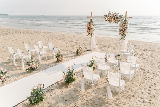 Beach Wedding Setup,a beautifully arranged setting for a wedding ceremony on a sandy beach. An elegant floral arch stands ready for the vows, flanked by rows of white chairs for the guests. 