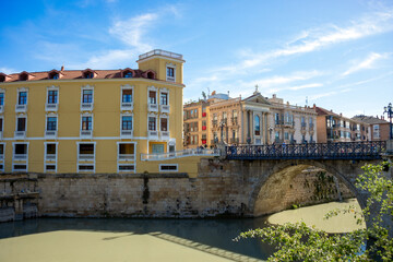 Puente de los Peligros over the Segura River, Murcia, Spain, with historic buildings in the background in bright afternoon light.