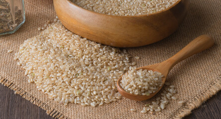 Background with brown rice grains in a wooden bowl and brown rice spread on a burlap cloth on a wooden table