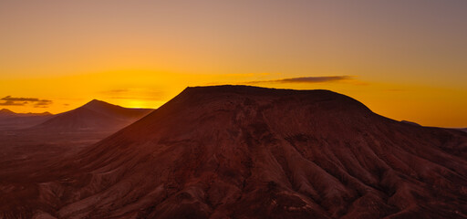 Beautiful aerial sunset image over Montana Roja volcanic crater silhouetted against the setting sun and skyscape near Corralejo, Fuerteventura, Canary Islands, Spain