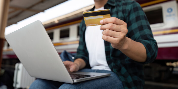 Travel and Finance. An excited traveler holding a credit card while working on his laptop at the train station.