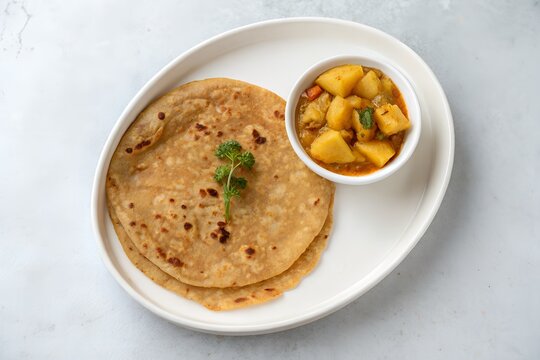 A single paratha with a small bowl of potato curry, top-down view, on a white ceramic plate, light grey background