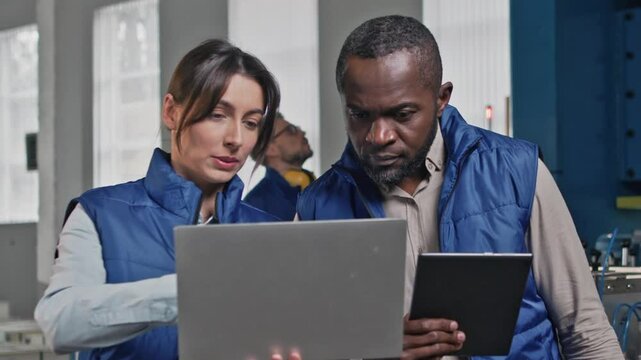 Concentrated female engineer holding laptop discussing data with male colleague using digital tablet. Collaborating on technical report or troubleshooting issue together inside production facility.
