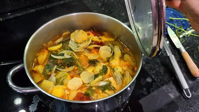 Traditional Portuguese codfish stew, known as caldeirada de bacalhau, prepared in a stainless steel pot with sliced potatoes, onions, tomatoes, peppers, herbs and olive oil.Homemade meal on a stovetop