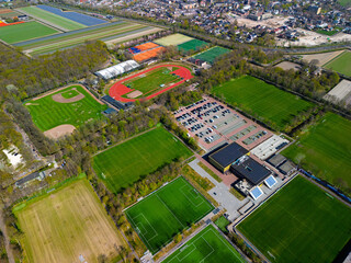 Aerial view of a sports complex with multiple football fields, a track and field stadium, tennis courts, and parking area, surrounded by lush forest and colorful tulip fields in the distance.